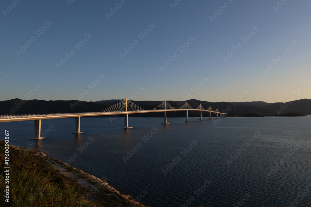 Obraz premium Scenic view of the modern Peljesac Bridge in Croatia spanning the calm Adriatic Sea during golden hour against dark mountains.