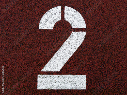 White number two painted on red running track surface, close-up of athletics field marking with textured background symbolizing progress, ranking, second place, and sports competition