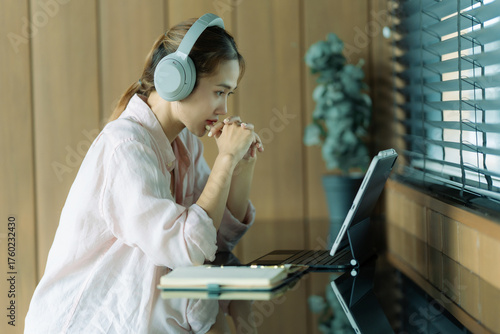 Smiling Asian female student wearing headphones for online learning using AI, listening to lectures, watching webinars, writing notes, and sitting in a coffee shop with a tablet.