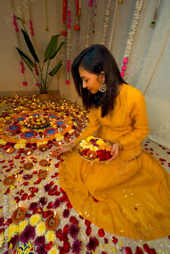 Happy young Indian woman wearing traditional yellow clothes holding plate with oil lamps and flowers celebrating Diwali festival. Selective focus. Festival of lights. Deepawali.