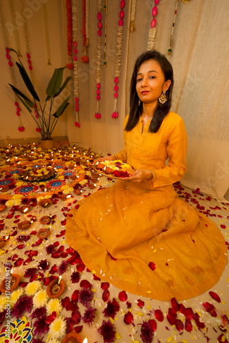 Happy young Indian woman wearing traditional yellow clothes holding plate with oil lamps and flowers celebrating Diwali festival. Selective focus. Festival of lights. Deepawali.