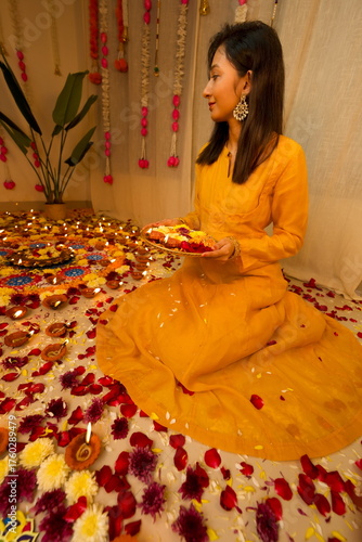 Happy young Indian woman wearing traditional yellow clothes holding plate with oil lamps and flowers celebrating Diwali festival. Selective focus. Festival of lights. Deepawali.
