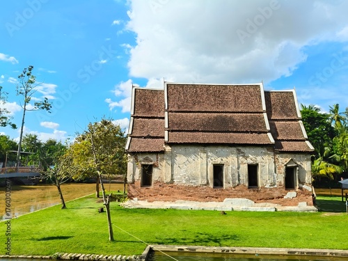 Historic Thai Temple Building: Old Architecture with Exposed Bricks, Aged Roof Tiles, and Peeling Stucco. Scenic View of Green Lawn and Water under Blue Sky.