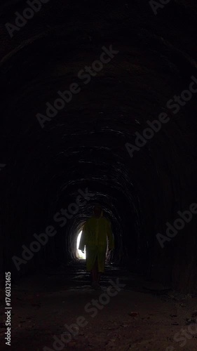 Vertical video. Woman in Yellow Raincoat Walks from Bright Tunnel Entrance into Deep Darkness Symbolizing Journey Through Fear and Self-Reflection Concept