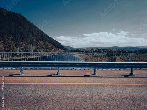 A view from a car, looking toward a bridge over a river, a small country road surrounded by fields, forests, and mountain ranges on the horizon. Cinematic blue depth effect.