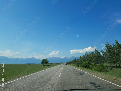 A view from a car onto a small country road with smooth asphalt, surrounded by fields, forests, and mountains on the horizon. Cinematic blue depth effect.