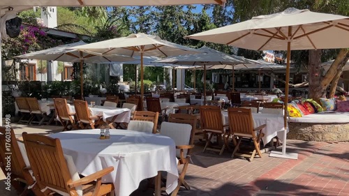 An elegant outdoor restaurant terrace features neat rows of wooden tables with white cloths and chairs, shaded by large umbrellas on a sunny day, perfect for dining and relaxation.
