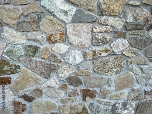 A detailed, full-frame shot showcasing the rugged, irregular texture of a stone wall constructed with various sizes of natural, multi-toned fieldstones and visible mortar joints.
