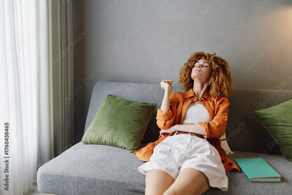 Naklejka premium Young woman in casual clothes relaxing on a sofa, exuding calmness and contemplation, with a stylish interior and natural light filtering through