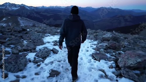 Person walks on a snowy, rocky trail toward the distant mountains at dusk, a cold landscape surrounds, a solitary figure with dark clothing and a hat hikes, the viewer observes from a rear...
