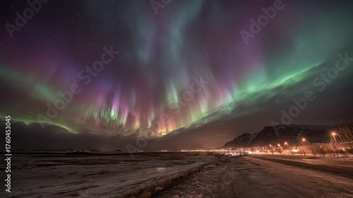 Wallpaper Mural Colorful northern lights fill the night sky with shades of green purple and blue above a serene landscape featuring mountains and a small town illuminated by streetlights. Torontodigital.ca