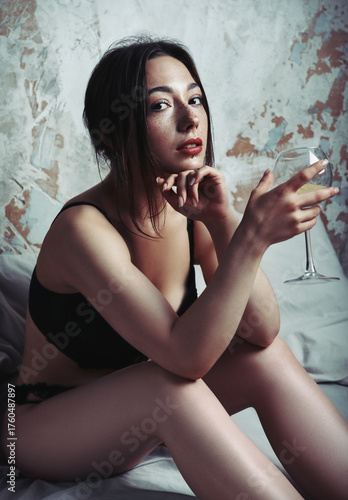 Portrait of lovely sad girl in black lingerie in old room. Beautiful young woman sitting on bed with white wine glass in hand