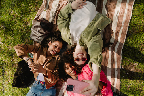 A group of three female friends are lying on a blanket and grass and smiling while posing in front of a phone that one of them is holding