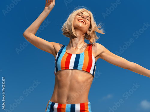 Joyful woman in colorful striped sportswear dancing under clear blue sky, expressing happiness, freedom, and summertime energy with a bright and vibrant atmosphere.