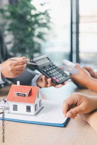 Close up of business people discussing mortgage loan with calculator, house model and contract paperwork on desk, concept of real estate finance, home ownership and property investment planning.