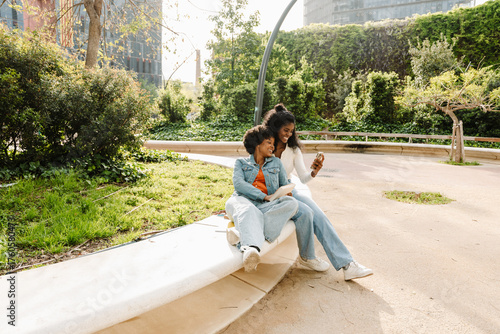 A female student holds a book and leans on a female student sitting next to her on a stone bench while they laugh and look at the phone one of them is showing