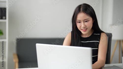 Young professional asian woman sitting at her desk, working on a laptop computer, smiling and reacting positively to something she sees on the screen, showing happiness and success