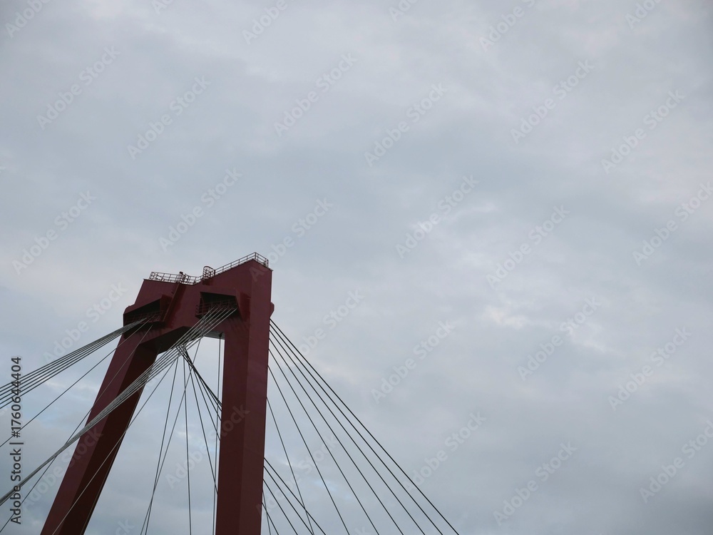 Fototapeta premium Red suspension bridge tower against dramatic cloudy sky, architectural close-up with steel cables. Engineering marvel, iconic structure, urban infrastructure, minimal color contrast.