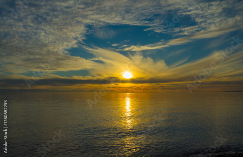 Issyk-Kul Lake at sunset, where the sun's golden glow meets wispy clouds and tranquil waters, creating a reflective symmetry under the vast morning sky, serene in its natural splendor.