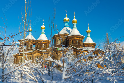 The venerable orthodox Holy Trinity Church in Karakol stands adorned with golden domes and crosses, its wooden frame contrasted by the white snow and the clear blue winter sky