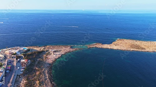 Aerial coastal peninsula view — narrow peninsula and adjacent harbor infrastructure, deep-blue calm sea with lighter nearshore shallows, scattered distant boats, and built-up shoreline.