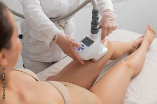 Caucasian girl receives cavitation vacuum roller massage on legs in beauty clinic. Woman cosmetologist performs LPG procedure to improve skin tone, reduce cellulite and sculpt lower body contours