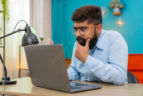 Canvas Print Indian man at home types on laptop stops rubs chin thinking over vital idea creative future plan