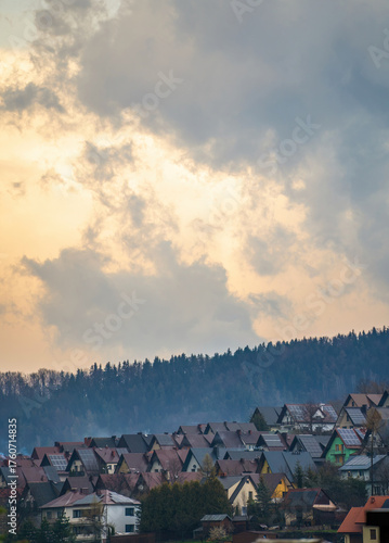 Fototapeta Naklejka Na Ścianę i Meble -  Charming mountain townscape in Limanowa, Poland, with colorful houses nestled below forested hills under a glowing sunrise or sunset sky.