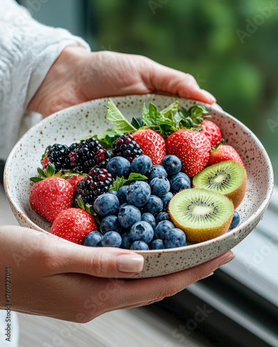 Fresh strawberries, blueberries, blackberries, and kiwi are beautifully arranged in speckled bowl, held by hands. This vibrant fruit medley evokes sense of health and vitality