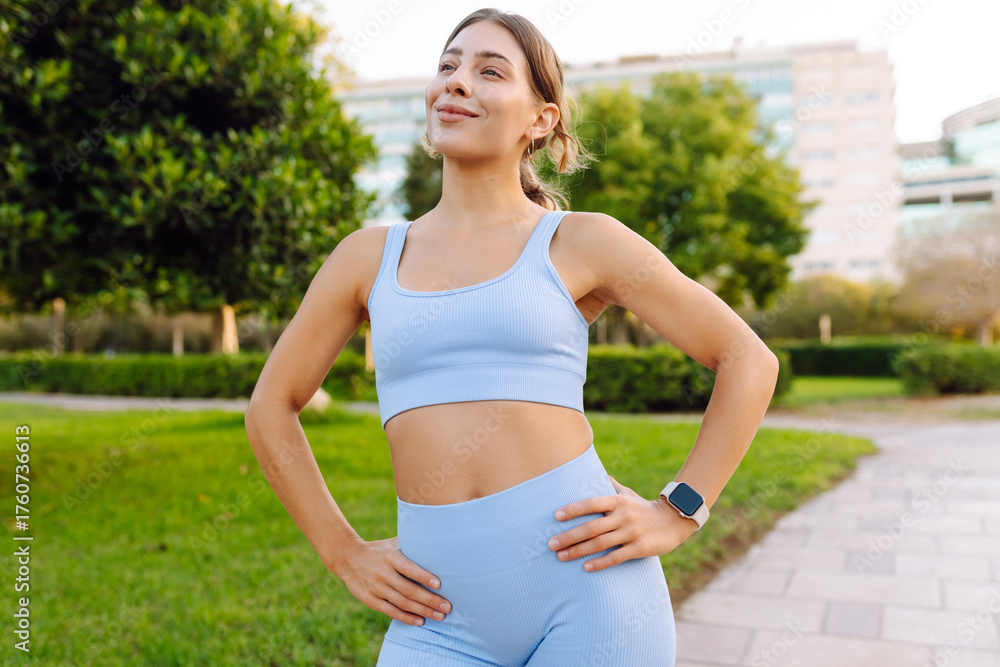 Fototapeta premium A young woman in athletic clothing enjoys a sunny day in a park. An athletic woman feels elated after a run and stretches outdoors. Concept of sport, enjoyment, and relaxation.