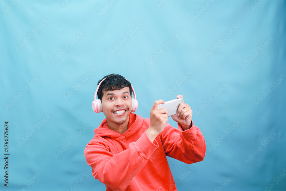 Naklejka premium Young man gaming with headphones and phone, smiling.