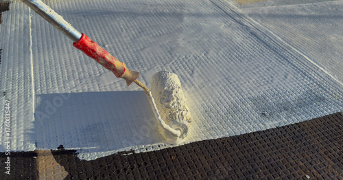 Worker applying white polyurethane waterproof coating on flat roof surface with paint roller.