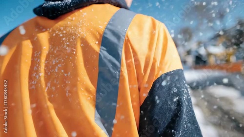 Close-up perspective from behind worker, orange jacket prominent, snow being cleared from sidewalk, winter snowfall creating soft blur in background