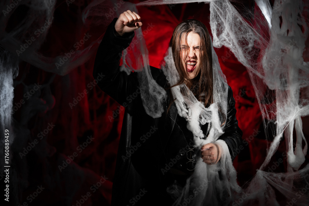 Obraz premium gothic woman with creepy vampire makeup trapped in spider web trying to scare showing strike gesture and fist-up. Fantasy scary girl on red halloween background.