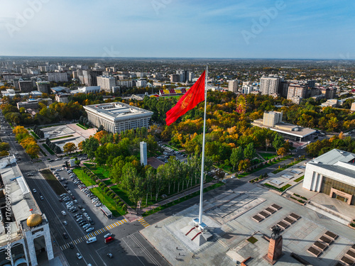 Aerial view of the large red Kyrgyz national flag on a flagpole over Ala-Too Square, surrounded by city buildings and autumn trees
