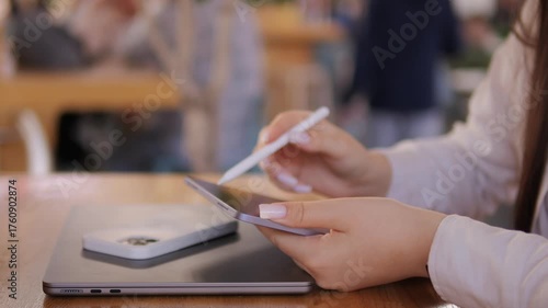 Woman is holding a pen and a tablet. She is sitting at a table. There are other people in the background