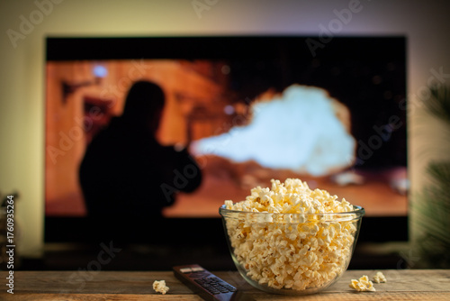 A glass bowl of popcorn and remote control in the background the TV works. Evening cozy watching a movie or TV series at home