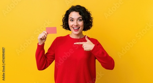 Cheerful woman with an excited expression holding and pointing to a blank credit card. Happy young person in a red sweater isolated on a yellow background with copy space