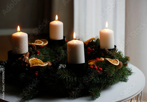Handmade advent wreath with four burning white candles, spruce branches, dried orange slices, cinnamon sticks and red berries, creating a warm and cozy Christmas atmosphere on a winter table