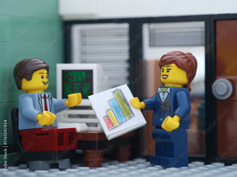 Naklejka premium Tambov, Russian Federation - October 19, 2025 A Happy Lego businessperson minifigure sitting behind a computer and talking with another Lego businessperson minifigure that is holding graphs.