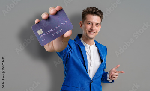 A young man stands with a smile, holding a purple card in one hand while his other hand points to it. He is dressed in a sharp blue suit, exuding confidence in his stance