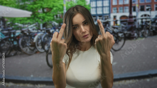 Young hispanic woman in white t-shirt flips middle fingers with bare hands on city street; anger defiance.