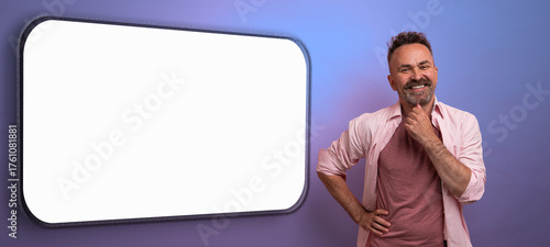 A joyful man stands near a blank sign in a colorful studio. He rests his hand on his chin, deep in thought, ready to share exciting ideas or messages on a vibrant purple backdrop