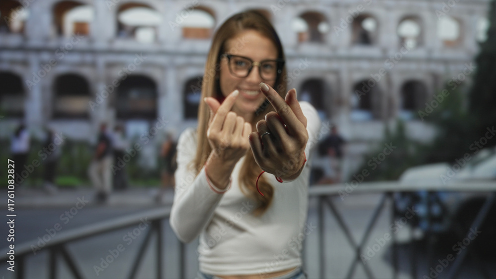 Fototapeta premium Woman making finger heart gesture with fingers and smiling in front of the roman coliseum building, casual outfit visible; playful travel.