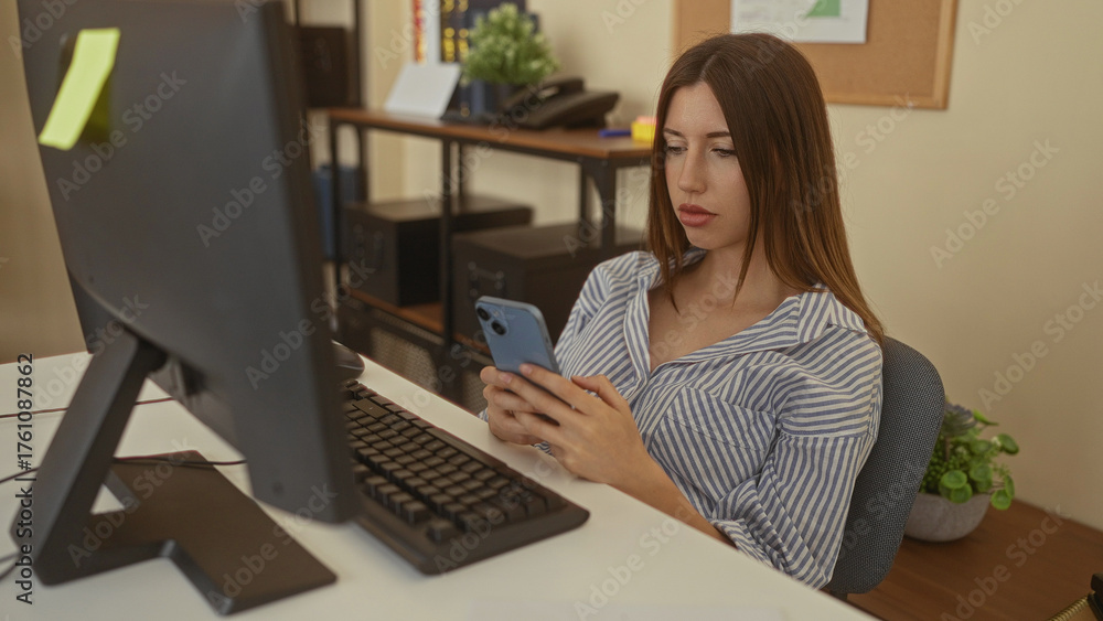 Fototapeta premium Woman holds smartphone and taps screen with hand in office building beside computer monitor; digital concentration.