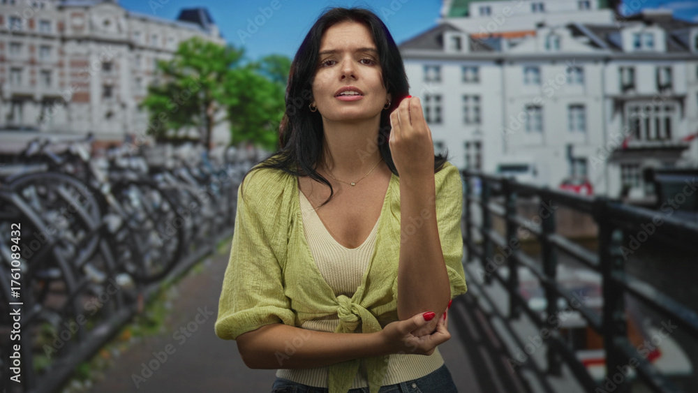 Fototapeta premium Young woman wearing a green blouse gestures with her hand and arm beside a canal on a city street; communication expression persuasion confidence.