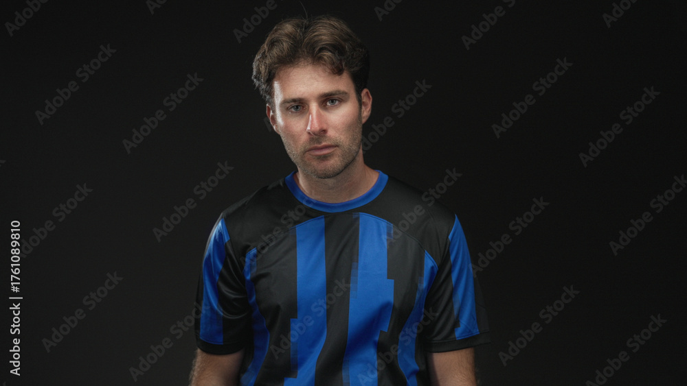 Naklejka premium Man smiling with slight head tilt wearing blue and black soccer jersey in studio against black backdrop; confidence.