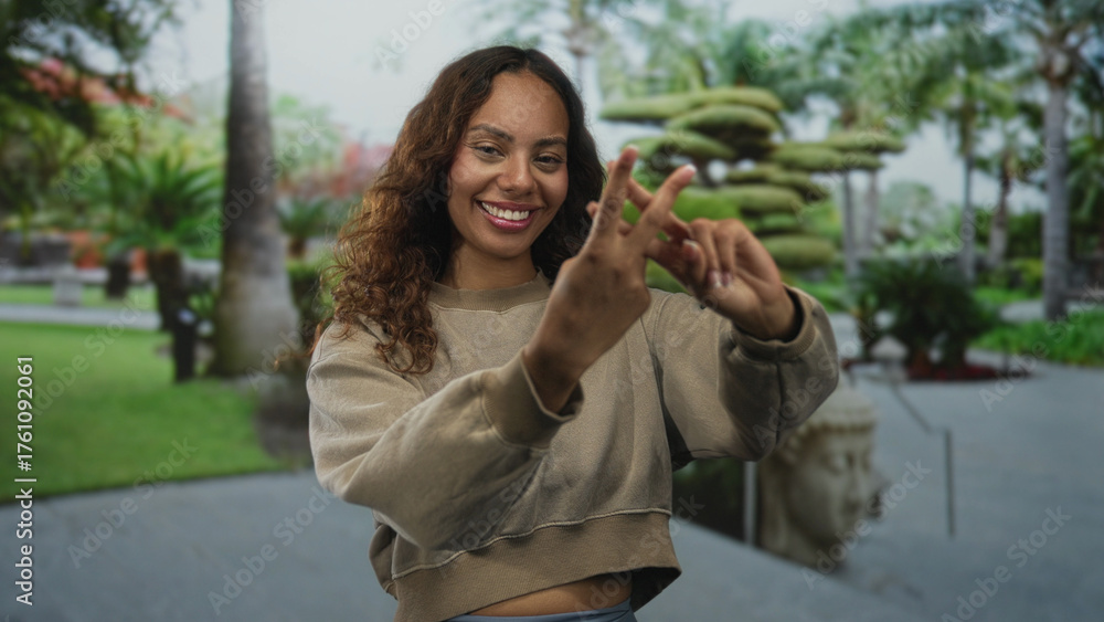 Fototapeta premium Smiling woman forms hashtag with fingers on tree lined street, bare hands in focus and casual cropped sweater; joy connection.