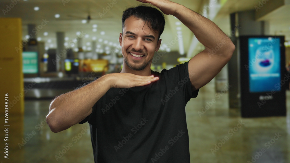 Fototapeta premium Young hispanic man smiling inside airport terminal with hands framing face conveying joy and excitement in modern indoor travel environment.