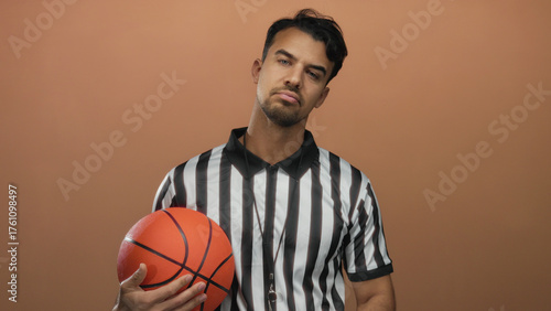 Young hispanic man in referee uniform holding basketball on brown background appears pensive and thoughtful, isolated with a serious expression.